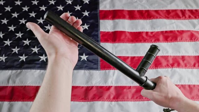 caucasian hands holding black rubber police tonfa baton blurry US flag. Someone menacingly pats himself on the hand with a nightstick.