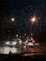 Night city view from the windscreen of the car at night. Rain drops on automobile window. Street lights reflections during summer shower. traveling by car in the evening in city town.
