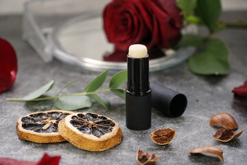 Fototapeta premium Homemade natural organic ingredients lip balm. Handmade lip balm and lipstick in black container on grey background and rose, eucalyptus, orange, nuts for decoration. 