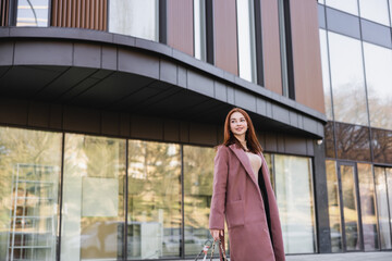 Fototapeta premium pleased redhead woman in coat standing near modern building.