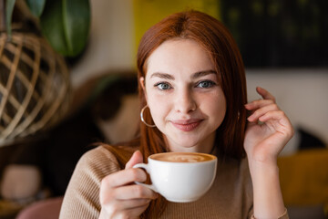 young redhead woman smiling and holding cup of latte in cafe.