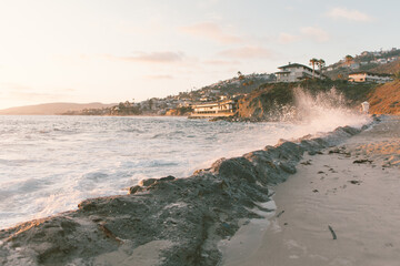 White waves crashing against beach rocks at sunset with building