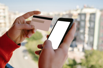 Young man wearing red hoodie holding smartphone with blank white screen and credit card or debit card, making financial transaction. Online payment. Mockup image of a hands holding credit card.