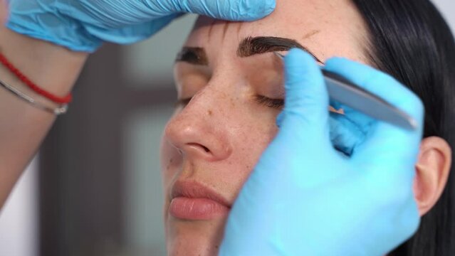 on a young female face, a master in gloves makes an eyebrow correction with tweezers, plucking out excess hairs