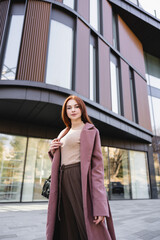 low angle view of redhead woman in coat standing with handbag near modern building.