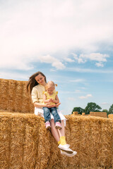 Naklejka na ściany i meble Portrait of young happy mother with little daughter in her arms sitting on haystack on farm. Hay harvesting.