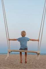 Blond boy swinging on the sand beach on sunset. Child swinging on seacoast. During the time sunrise and sunset.