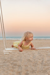 Cute fair-haired girl get on the swings on sandy beach. Little girl has fun by the sea during sunset.