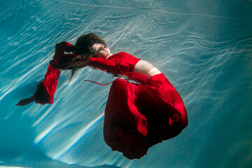 Attractive red-haired young woman swims beautifully underwater in a red dress