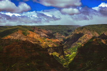 Fototapeta premium Waimea Canyon on the Hawaiian Island of Kauai, with red rocks and bright green foliage, dark gloomy clouds, bright blue sun, and long beautiful waterfalls 