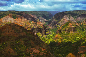 Fototapeta na wymiar Waimea Canyon on the Hawaiian Island of Kauai, with red rocks and bright green foliage, dark gloomy clouds, bright blue sun, and long beautiful waterfalls 