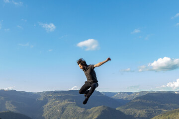 Young man dressed in black clothes jumping on mountains and blue sky background, in Nova Petropolis, Rio Grande do Sul sierra, Brazil - obrazy, fototapety, plakaty