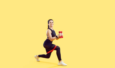 Athletic lady isolated on yellow enjoying fitness workout. Full length studio shot of happy beautiful fit woman in sportswear doing exercise with elastic band and dumbbells. Body training concept