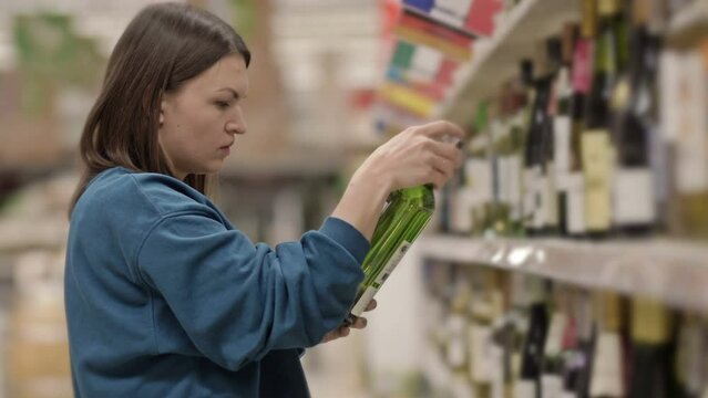 Young woman chooses wine while standing in front of shelves with alcohol in a supermarket or liquor store.