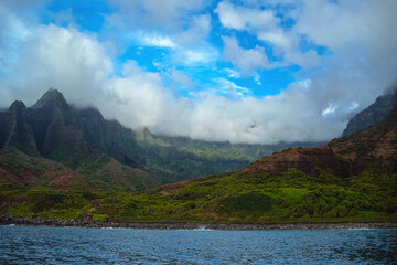 The gorgeous rugged wilderness and cliffs of Kauai's Napali Coast in Hawaii, with low clouds and mist hanging over the mountain peaks under a stormy grey sky, and bright blue and teal ocean waves