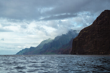 Naklejka premium The gorgeous rugged wilderness and cliffs of Kauai's Napali Coast in Hawaii, with low clouds and mist hanging over the mountain peaks under a stormy grey sky, and bright blue and teal ocean waves