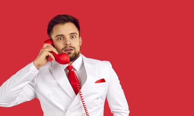Shocked man holding handset and listening to unbelievable news on red background. Young man in...