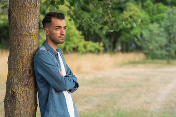 .A sad young man with stubble in a denim shirt stands under a tree