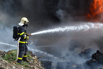 A fireman extinguishes huge a landfill fire with flames and black smoke in the background