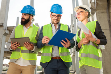 Happy mixed group of young architects singing building progress reports and talking during meeting on a construction site