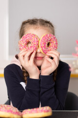 Curious child with braided hair holding up two sprinkled donuts to her eyes in a modern kitchen setting.