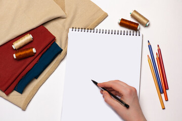 Mock up of white notebook and hand with pen on it. Sewing threads, fabric and pencils on white background.