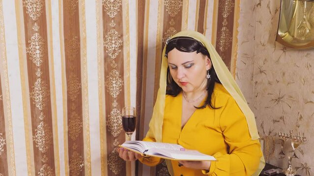 A Jewish woman in a wig and head covering makes a celebratory kdush with a glass in her hand and a siddur.