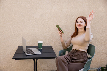 redhead freelancer in turtleneck holding smartphone while calling waiter on summer terrace.