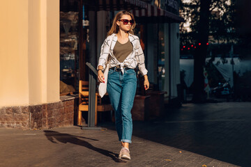 Cool girl with shopper bag walks on a sunny city street in sunglasses and casual clothing. The sun shines on the girl. Sunny day in the city.