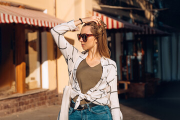 Cool girl walks on a sunny city street and holding a hairstyle in sunglasses and casual clothing. The sun shines on the girl. Sunny day in the city.