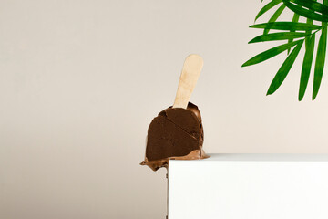 Broken chocolate ice cream bar with stick on white podium and palm branch on pastel color background. White platform with chocolate popsicle. Summer creative concept with copy space.