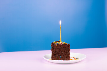 Chocolate cake slice with candle on pink and shine golden background. Birthday party minimal...