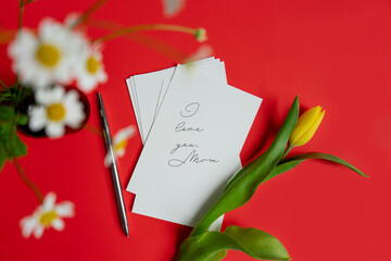 Obraz premium Mother's Day message with flowers and candies. Congratulations on mother's day with a card and spring flowers yellow tulip on a red flat lay background.