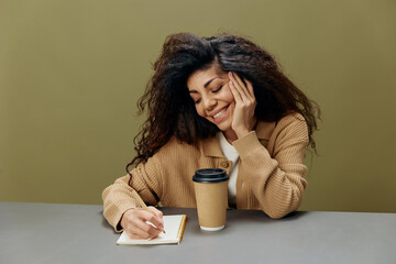 Enjoyed smiling cute tanned curly Latin lady recline on hand write essay letter sit at the table isolated over olive green background look at notebook. Copy space Mockup Banner. Distance education