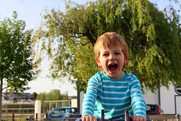 A blond boy with a wide open mouth on a blurred green background. Emotional five-year-old boy on the playground.