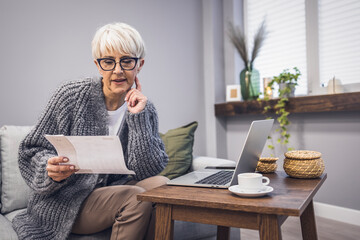 Serious aged woman in eyeglasses checking all bills, calculating expenses. Mature housewife sitting at table with laptop, looking through financial papers with focused expression.  - obrazy, fototapety, plakaty