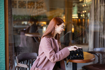 young and happy woman with red hair looking at handbag in cafe terrace.