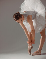 classic ballet posing. young slim dancer, brunette girl ballerina is tying ribbons on pointe shoes...