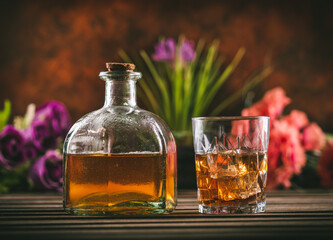 Glass of whiskey with ice and bottle with liquor on a wooden table with some color flowers. Alcoholic beverage ready to drink.