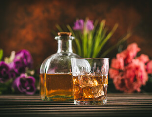 Obraz na płótnie Canvas Glass of whiskey with ice and bottle with liquor on a wooden table with some color flowers. Alcoholic beverage ready to drink.