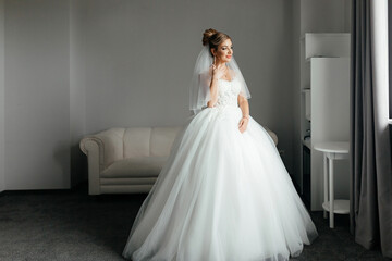 Fototapeta premium Bride morning concept. An elegant bride stands at the window of a hotel room and looks into the camera.