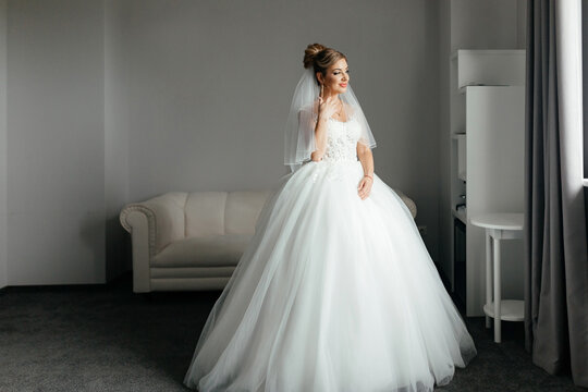  Bride morning concept. An elegant bride stands at the window of a hotel room and looks into the camera.