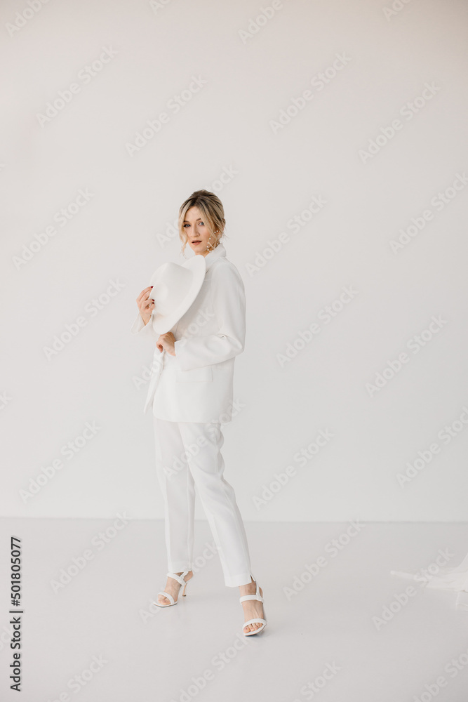 Wall mural Confident young blond woman smiling, looking at camera isolated on white background. Studio portrait of successful friendly female in white suit and hat, posing over white wall.