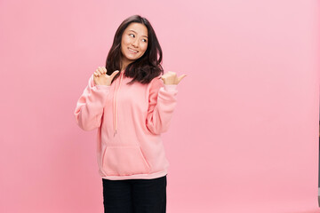 Lovely cheerful Asian student young lady in pink hoodie sweatshirt point finger aside at copy space for ad posing isolated on over pink studio background. Good offer. Fashion New Collection concept