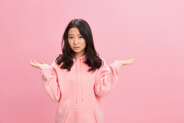 Pensive lovely Asian student young lady in pink hoodie sweatshirt spreading hands aside posing isolated on over pink studio background. Good offer. Fashion New Collection concept