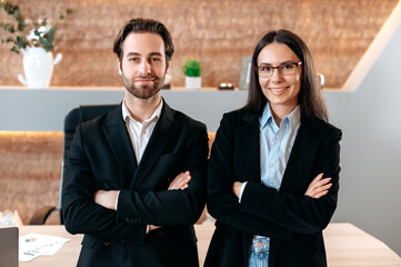 Portrait of two successful caucasian business partners, creative man and woman, corporate employees in a modern office, dressed in formal wear, looking at the camera, with their arms crossed,smile