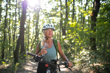 Fototapeta na wymiar Happy senior woman biker drinking water from bottle outdoors in forest.