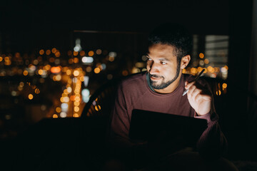 Young African American man sitting on balcony with urban view and using tablet at night