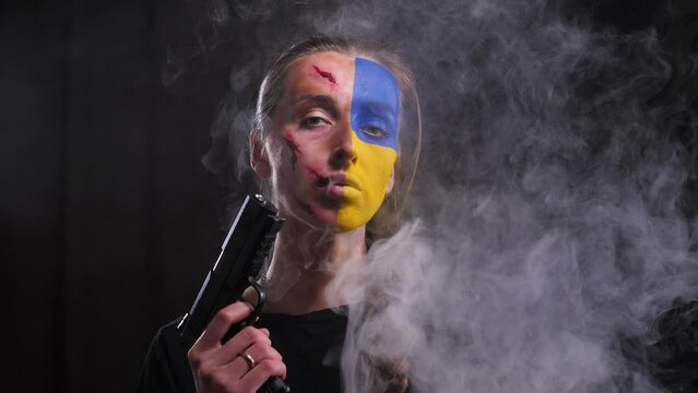 a woman with wounds and the flag of Ukraine on her face holds a gun in her hand turns and looks at the camera