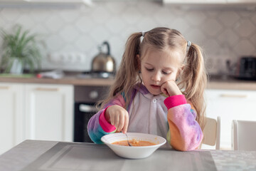 cute little girl eats soup at home in the kitchen without appetite. The child sadly looks at the plate, does not want to eat. Healthy food, natural vitamins, baby food concept. selective focus